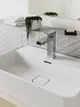 Edelstahl-Waschtischarmatur von Ideal Standard, montiert auf einem weißen Becken, mit quadratischem Ablauf und minimalistischer Formgebung. Handtücher und Pflanze im Hintergrund sichtbar.